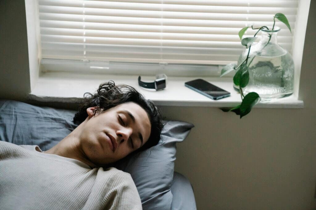 High angle of young ethnic male sleeping in bed with smartphone and smart watch placed near head on windowsill